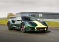 A green Lotus Emira Jim Clark Edition with a yellow stripe on its hood is parked on a racetrack in front of a modern building.