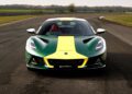 A green Lotus Emira Jim Clark Edition sports car with a yellow racing stripe is parked on a racetrack, surrounded by grass and trees in the background.