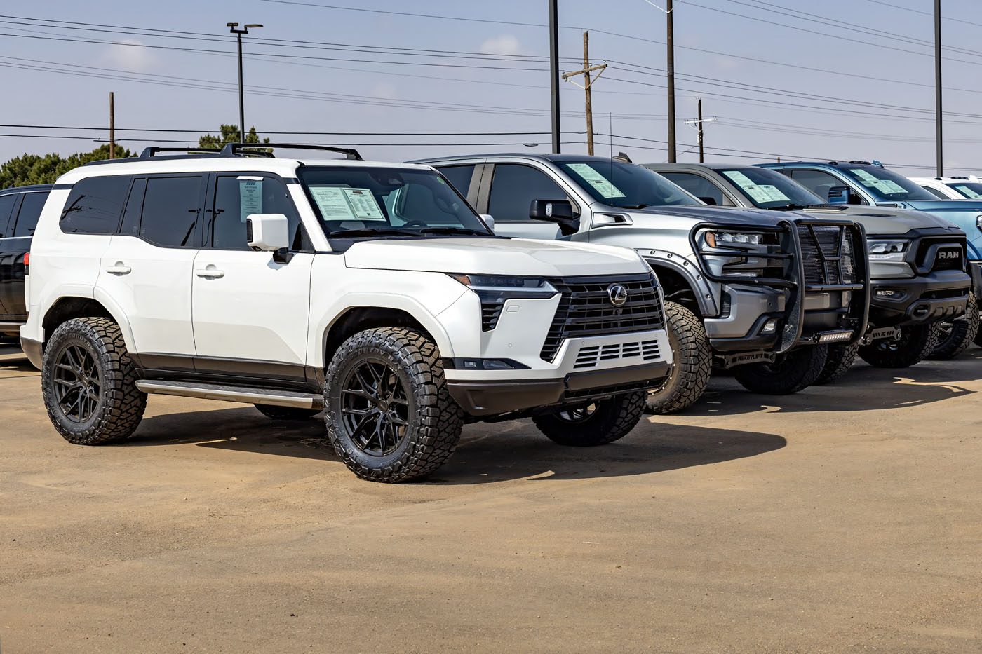 A row of large SUVs and trucks, including a white Lexus SUV with off-road tires, sits on a dealership lot under a clear sky—perfect for anyone searching for lifted trucks in America.
