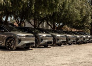 A row of identical silver electric cars is parked side by side on a gravel lot under large trees.
