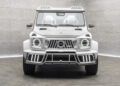 A white, customized Mansory Mercedes-Benz G-Class SUV viewed from the front, featuring a unique grille, headlights, roof lights, and sporty body modifications, parked in a gray showroom.