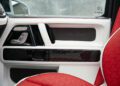 Close-up of a luxury G-Class door interior featuring white and black accents, red stitching, control buttons, "Mansory" branding, and part of a red and white seat.