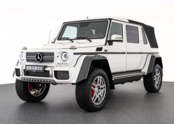 A unique creation, this white Brabus-modified Mercedes-Benz G-Class SUV with large off-road tires and a black soft-top roof is parked indoors on a gray garage floor.