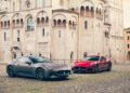 Two Maserati GranTurismo sports cars, one gray and one red, are parked on a cobblestone square in front of an ornate stone Modena building.