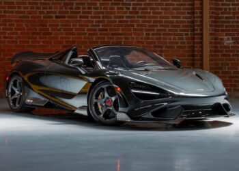 A black McLaren 720S Spider with the roof down is parked indoors in front of a brick wall, exuding the racing heritage seen in icons like the LM25 and the McLaren 765LT Spider.