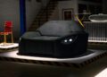 A black sofa designed to resemble the front of a car, complete with headlight-shaped lights and inspired by Mercedes-AMG performance car parts, is displayed on a platform in an indoor setting.