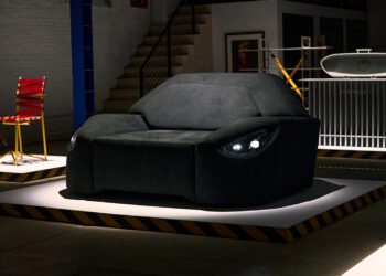 A black sofa designed to resemble the front of a car, complete with headlight-shaped lights and inspired by Mercedes-AMG performance car parts, is displayed on a platform in an indoor setting.