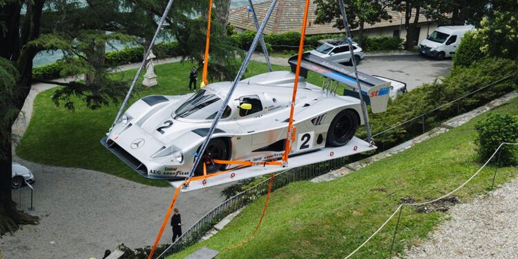 A silver Mercedes-Benz CLK GTR race car is being lifted by orange straps and a crane over a grassy hill, near a driveway with trees and parked vehicles in the background, possibly for FuoriConcorso 2025.