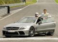 A man in a racing suit stands by the open driver's door of a silver Mercedes-Benz sports car—the only Mercedes-AMG Black Series to feature a V12—parked on a racetrack.
