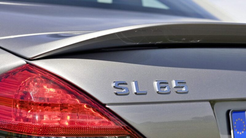 Close-up of the rear of a silver car showing the taillight, a raised spoiler, and the badge "SL 65"—the Only Mercedes-AMG Black Series To Feature A V12—on the trunk.