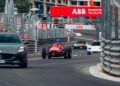 A vintage red race car leads modern sports cars around a bend on a closed street circuit, with an ABB sign visible overhead.