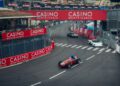 Four race cars speed around a curve on the city track at the Monaco E-Prix, passing under red "Casino Monte-Carlo" banners, with buildings and fencing in the background.