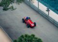 A red vintage Maserati race car with the number 30, driven by a person in a blue helmet, travels on a road beside a body of water and a metal barrier, evoking the thrilling spirit of the Monaco E-Prix.