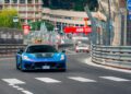 A blue Maserati drives on a racetrack with barriers during the Monaco E-Prix, followed by another car; city buildings and race signage are visible in the background.