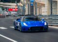 A blue Maserati supercar speeds along a fenced street circuit at the Monaco E-Prix, with barriers and signage visible in the background.