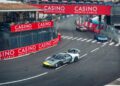 Several sports cars race around a bend on a city street circuit during the Monaco E-Prix, passing under red Monte-Carlo Casino banners.