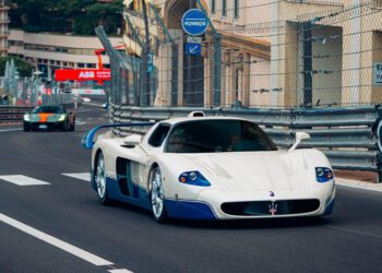 A white and blue Maserati MC12 supercar speeds along a fenced city racetrack during the Monaco E-Prix, with another sports car close behind.