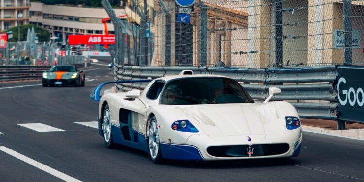 A white and blue Maserati MC12 supercar speeds along a fenced city racetrack during the Monaco E-Prix, with another sports car close behind.