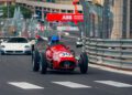 A vintage red race car with the number 30 leads the Monaco E-Prix Supercar Parade on a racetrack, followed by Maserati and other modern sports cars, with barriers and fencing lining the circuit.