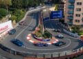 Six sports cars, including a Maserati, race around a tight hairpin turn on a city street circuit during the Monaco E-Prix, surrounded by barriers, plants, buildings, and "Monaco" signs and advertisements.