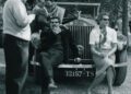 A group of people are gathered around a vintage Rolls-Royce Phantom parked on a gravel path, with two individuals sitting on the front bumper and others standing nearby in a wooded area, reminiscent of gatherings by celebrity owners.