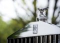 Close-up of the iconic Rolls-Royce hood ornament and logo on a Phantom’s front grille, with blurred greenery in the background—a favorite among celebrity owners.