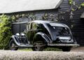 A vintage black Rolls-Royce Phantom with chrome accents is parked on gravel before a rustic wooden building and lush greenery, evoking the timeless style favored by celebrity owners.