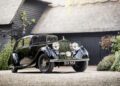 A vintage black Rolls-Royce Phantom with the license plate DUV 553 is parked on a gravel driveway before a rustic, thatched-roof building—echoing the elegance often favored by celebrity owners.