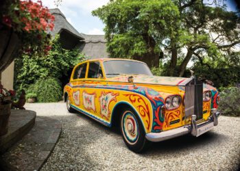 A classic Rolls-Royce Phantom is parked in a gravel driveway, featuring a colorful, psychedelic paint design with intricate patterns and motifs reminiscent of its famous celebrity owners.