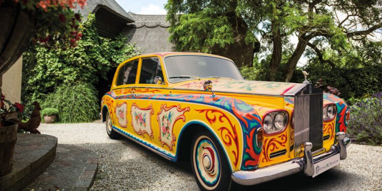 A classic Rolls-Royce Phantom is parked in a gravel driveway, featuring a colorful, psychedelic paint design with intricate patterns and motifs reminiscent of its famous celebrity owners.