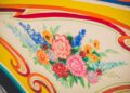 A colorful floral painting featuring red, blue, yellow, and orange flowers with green leaves on a cream background with swirling decorative borders—an art piece fit for celebrity owners or the interior of a luxurious Rolls-Royce Phantom.