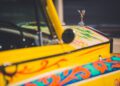 Close-up of a vintage yellow Rolls-Royce Phantom with colorful, ornate designs on the hood and a silver winged hood ornament—an iconic style often favored by celebrity owners.