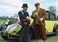 Two men stand beside a vintage yellow Rolls-Royce; one in a suit and top hat holds a golf bag, while the other in a cap and knickerbockers examines a golf club, recalling the style of early Phantom celebrity owners.