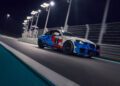 A BMW M2 with a blue and red camouflage pattern races on a nighttime track under bright lights, capturing the adrenaline of the 24h Nürburgring.