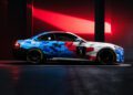 A sporty BMW M2 Racing car with a geometric blue, white, and red camouflage design and the number 1 on the door is parked indoors under red lighting, evoking the endurance spirit of the 24h Nürburgring.