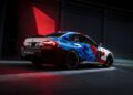 A BMW M2 Racing car with a blue, red, and white pixelated livery is parked indoors, illuminated by dramatic red and white lighting—ready for its next 24h Nürburgring challenge.