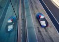 A BMW M2 Racing car with a blue, red, and white geometric design is parked on the pit lane next to a building during the 24h Nürburgring, its reflection visible in the glass.