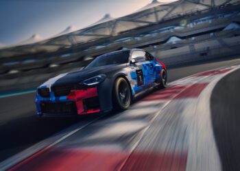A BMW M2 Racing car with a blue, black, and red livery speeds around a track curve during the 24h Nürburgring, with empty grandstands visible in the background.