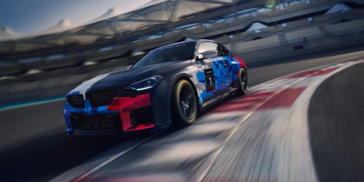 A BMW M2 Racing car with a blue, black, and red livery speeds around a track curve during the 24h Nürburgring, with empty grandstands visible in the background.