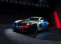 A BMW M2 Racing car with a pixelated blue, red, black, and gray livery is parked in a dark, industrial setting under spotlights, ready for the 24h Nürburgring.