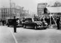 A black-and-white photo shows a Rolls-Royce Phantom motorcade stopped near a crowd and a canopy, with a uniformed officer saluting as people watch on a city street lined with historic buildings.