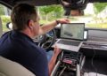 A man sits in the driver's seat of a BMW i7, using a laptop connected to the car's onboard computer system for diagnostics or data analysis, possibly checking performance data from its solid-state EV batteries.