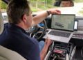 A man in a BMW car holds a laptop displaying technical data, seated next to exposed electronic equipment and discussing innovations like all-solid-state EV batteries in the center console.