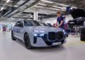 A person in a workshop inspects a silver BMW sedan with headlights on, possibly assessing the i7 or models featuring solid-state EV batteries; another car is raised on a lift in the background.