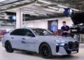 A person stands next to a silver BMW i7 test vehicle featuring solid-state EV batteries inside a brightly lit automotive workshop, with another car visible on a lift in the background.