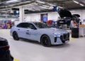 A silver BMW sedan with "BMW Group Hydrogen Fuel Cell Power" branding is parked in an auto workshop; in the background, another BMW—possibly an i7 being serviced for all-solid-state EV batteries—sits on a lift.