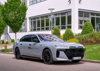 A silver BMW i7 electric sedan is parked by a curb, plugged in for charging, with a modern building and greenery in the background.
