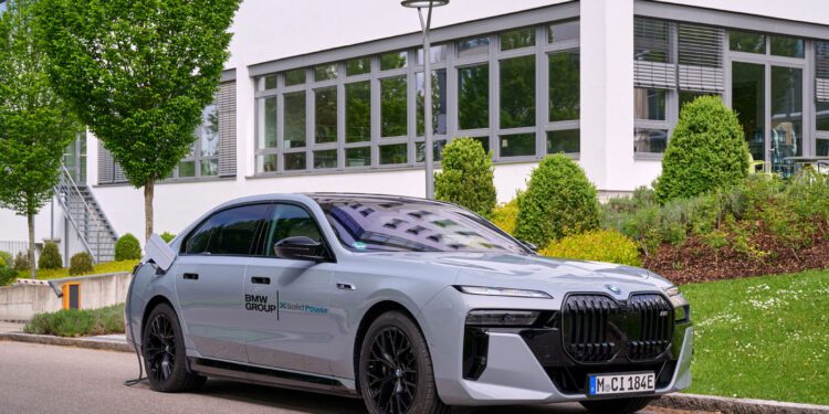 A silver BMW i7 electric sedan is parked by a curb, plugged in for charging, with a modern building and greenery in the background.