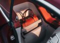 A leather handbag sits secured on the rear seat of a BMW with white and red interior, illuminated by sunlight through the open door.