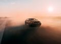 A black Speedtop V8 sports car drives on an open road at sunset, kicking up dust behind it under a clear sky.
